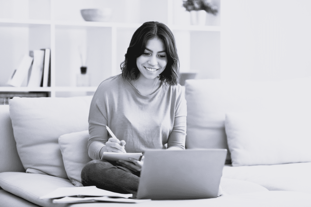 Ung pige sidder på sofa og skriver i notesblok og arbejder på bærbar